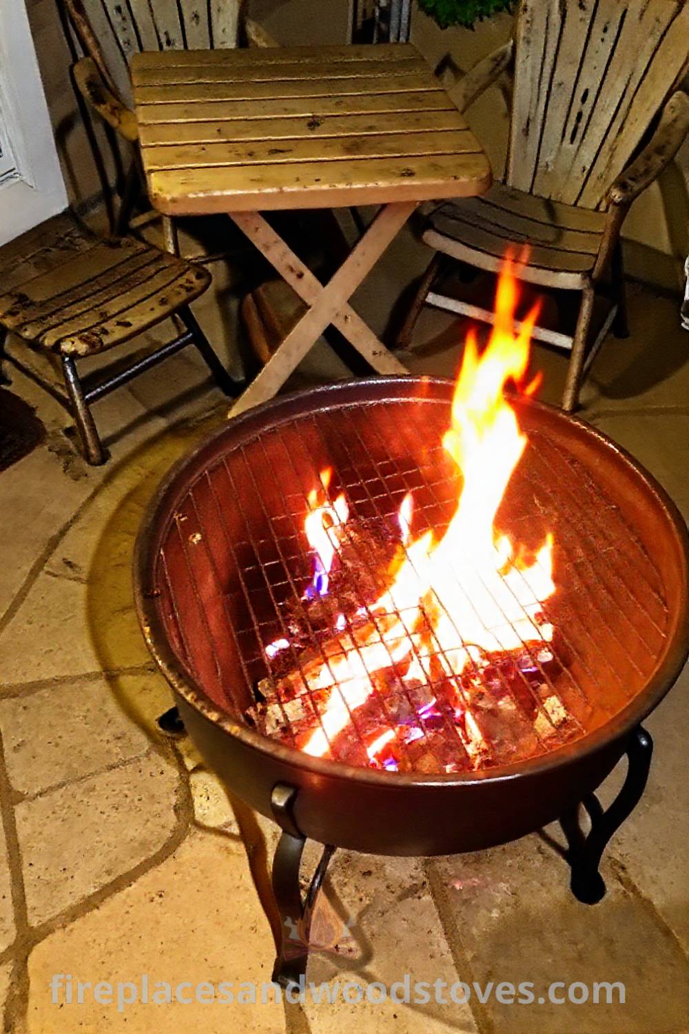 Metal charcoal-burning fire pit at the center of a cozy patio, surrounded by mismatched chairs and a wooden table, creating an inviting outdoor space for gatherings under the stars. Visit patioandhomefurniture.com for stylish designs transforming your backyard.