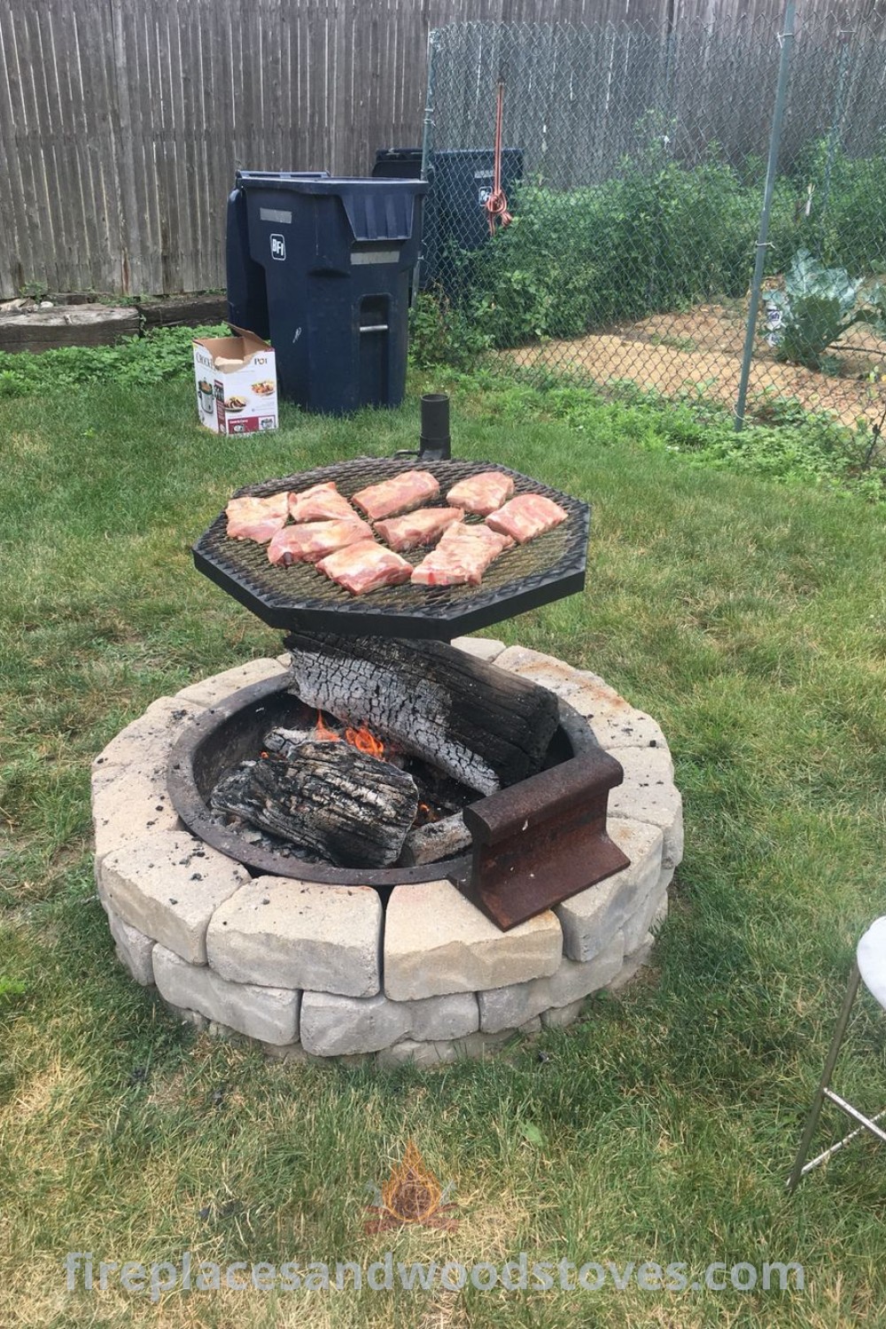 An outdoor BBQ grill with food on it in the grass next to a trash can, showcasing a stylish setup with an in-ground BBQ pit and a washing machine drum fire pit. Ideal for creating a cozy and functional space for grilling and cooking. Discover more outdoor ideas at patioandhomefurniture.com.