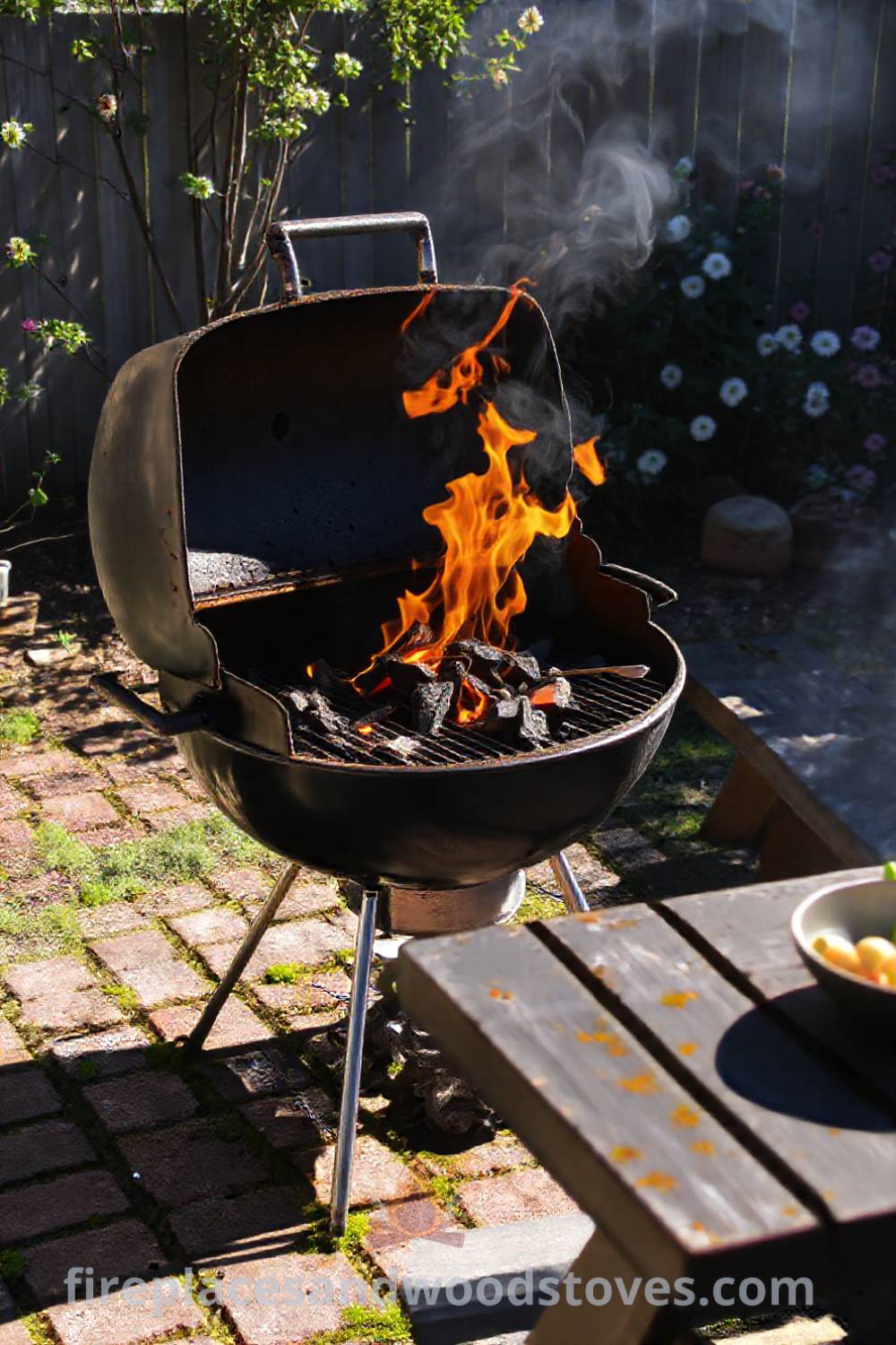 Charcoal grill barbecue in a sun-drenched backyard with warm flames, uneven brick patio, picnic table with fresh vegetables, and blooming flowers creating a cozy and inviting outdoor space. Visit patioandhomefurniture.com for incredible ideas that will wow your guests.