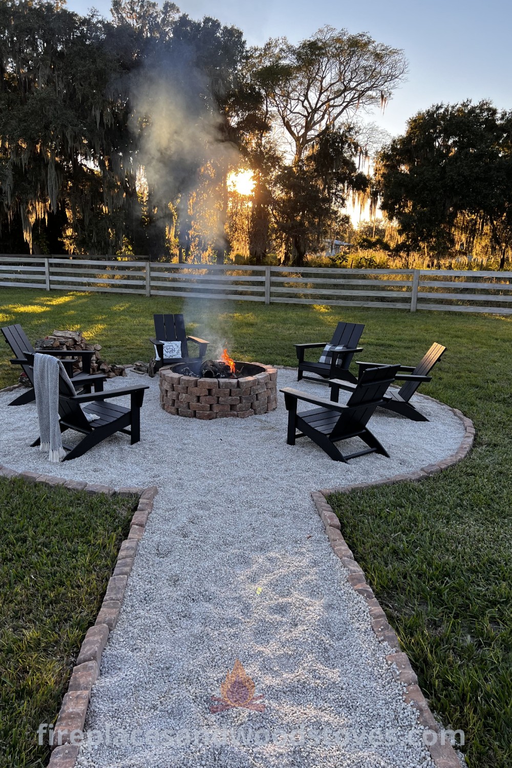 A cozy gravel fire pit area with chairs encircling it, set in a grassy landscape next to a fence, showcasing charming fire pit ideas from patioandhomefurniture.com.