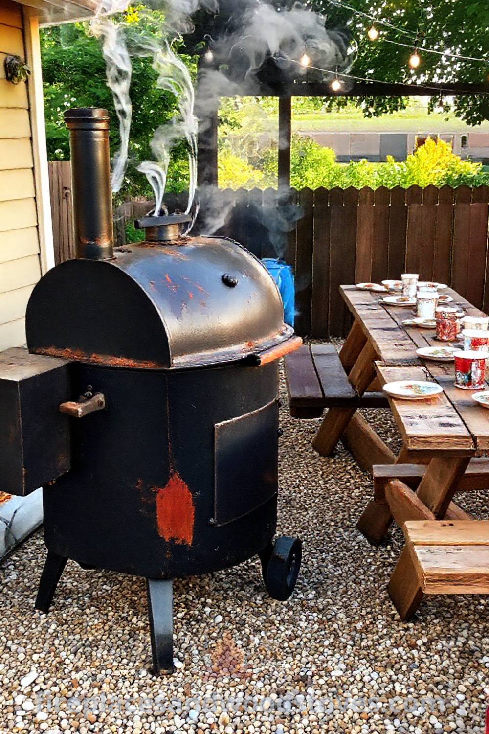 Outdoor BBQ smoker on a gravel patio surrounded by picnic tables with mismatched plates, creating a cozy gathering space filled with hickory aromas, perfect for summer cookouts. Visit patioandhomefurniture.com for incredible ideas transforming your backyard.