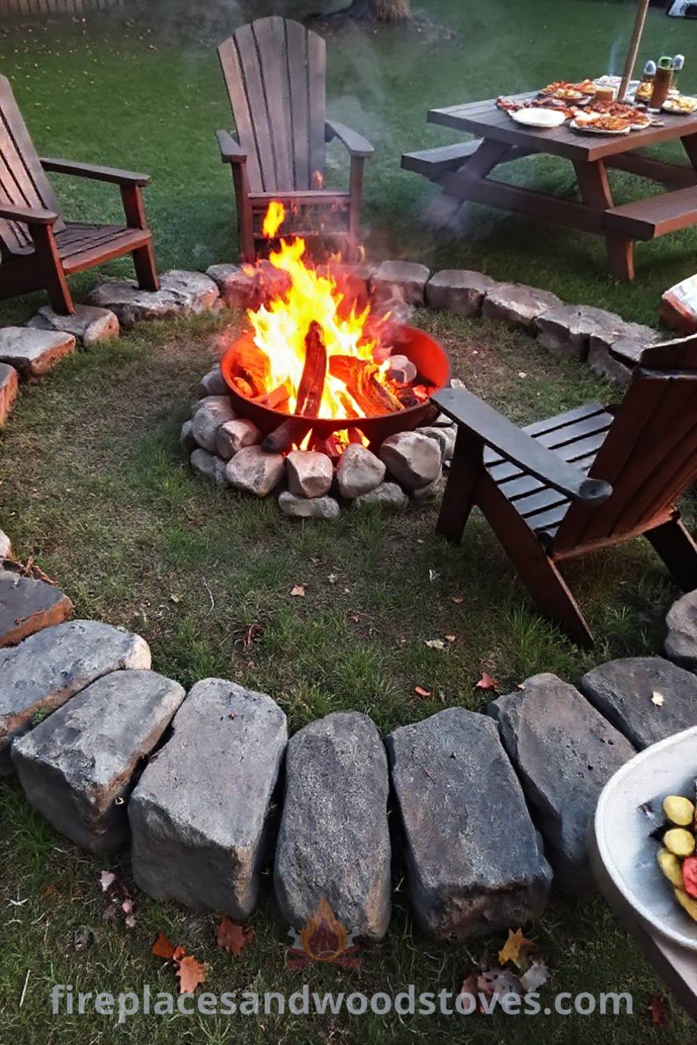 Cozy charcoal camping fire pit surrounded by uneven stones, illuminated by bright orange flames and complemented by a picnic table full of snacks for a warm gathering atmosphere. Visit patioandhomefurniture.com for incredible ideas transforming your backyard into the coziest home.