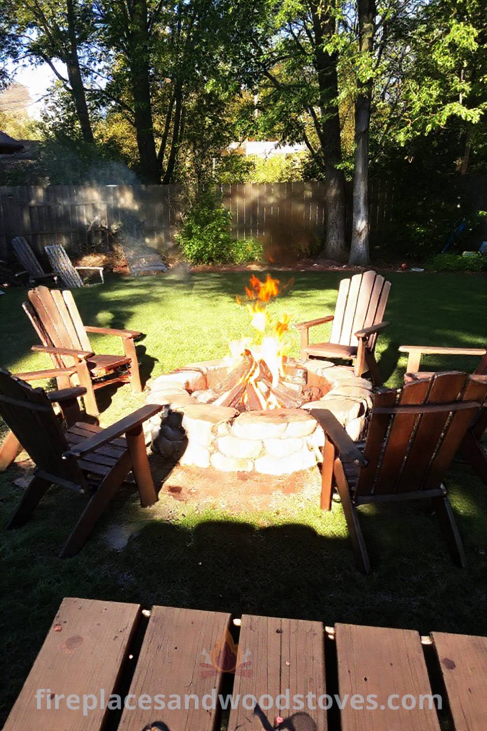 Stone fire pit surrounded by mismatched wooden chairs in a sun-dappled backyard, creating a cozy and inviting atmosphere for gatherings. Visit patioandhomefurniture.com for incredible ideas transforming your backyard into a stylish oasis.