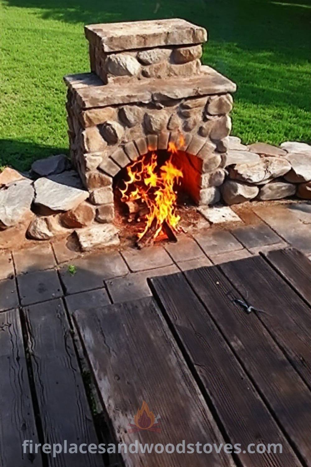 Stone fireplace BBQ in a grassy backyard surrounded by weathered wooden patio, with flames flickering and a picnic table nearby, creating a cozy and inviting atmosphere for gatherings. Visit patioandhomefurniture.com for stylish outdoor design inspiration.
