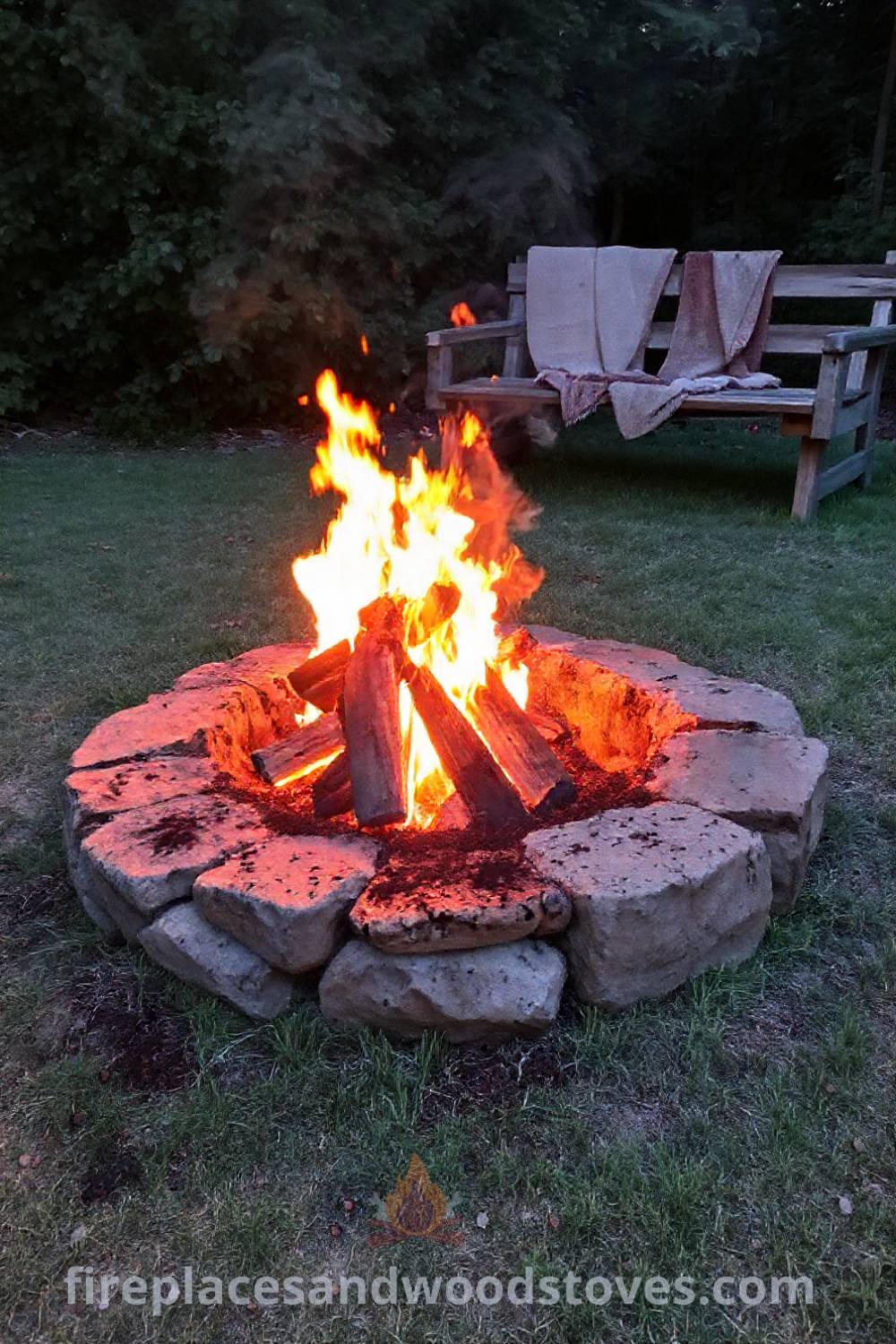 Rustic charcoal fire pit made of rough-hewn stone surrounded by mismatched wooden benches and cozy blankets, creating a warm and inviting outdoor gathering space perfect for evenings under the stars. Visit patioandhomefurniture.com for incredible ideas that will wow your guests.