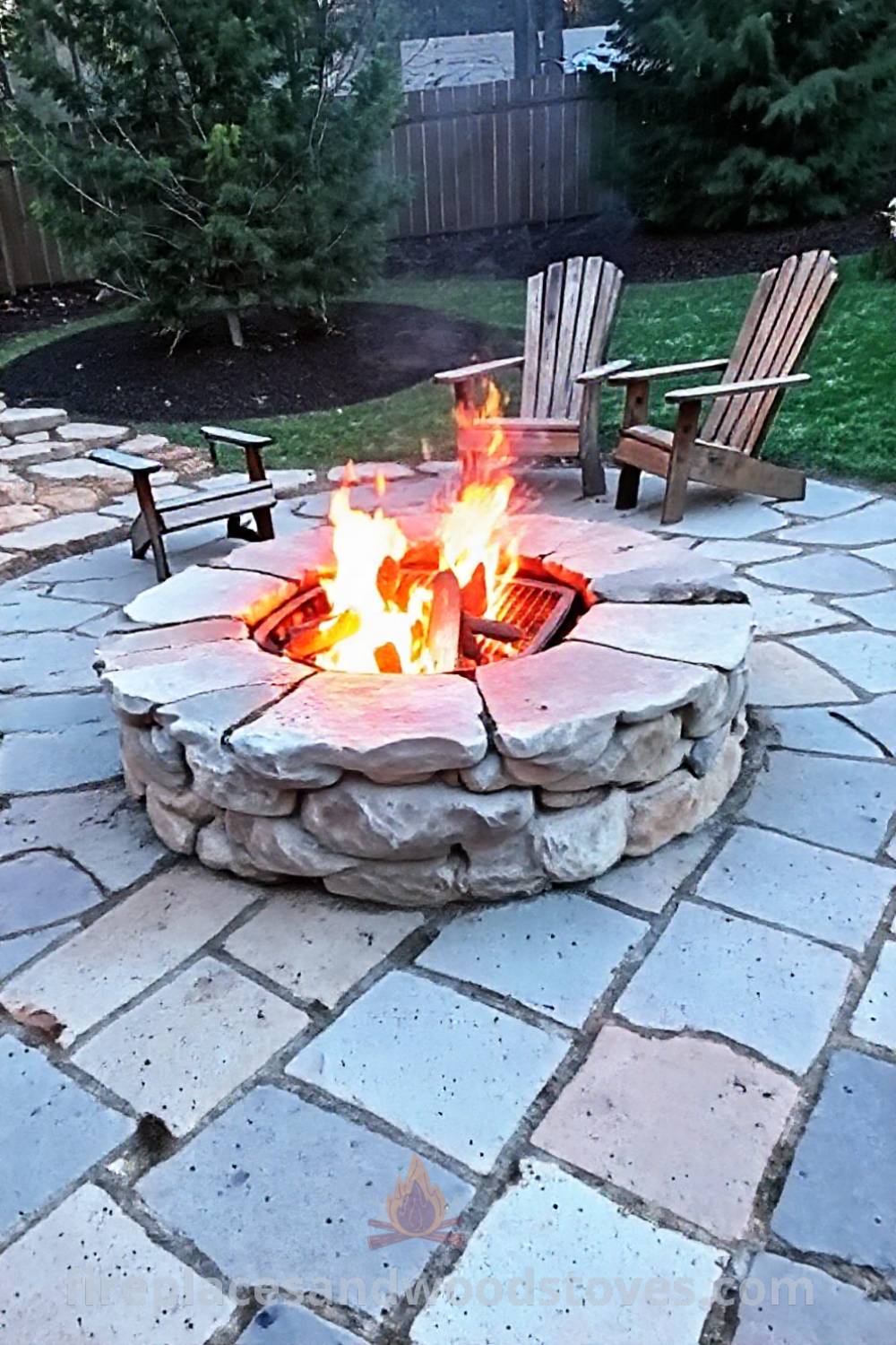 Backyard stone wood-burning fire pit with built-in grill surrounded by rustic chairs and uneven flagstones, creating a cozy gathering space ideal for sharing stories under the starlit sky. Visit patioandhomefurniture.com for stylish ideas that will wow your guests.