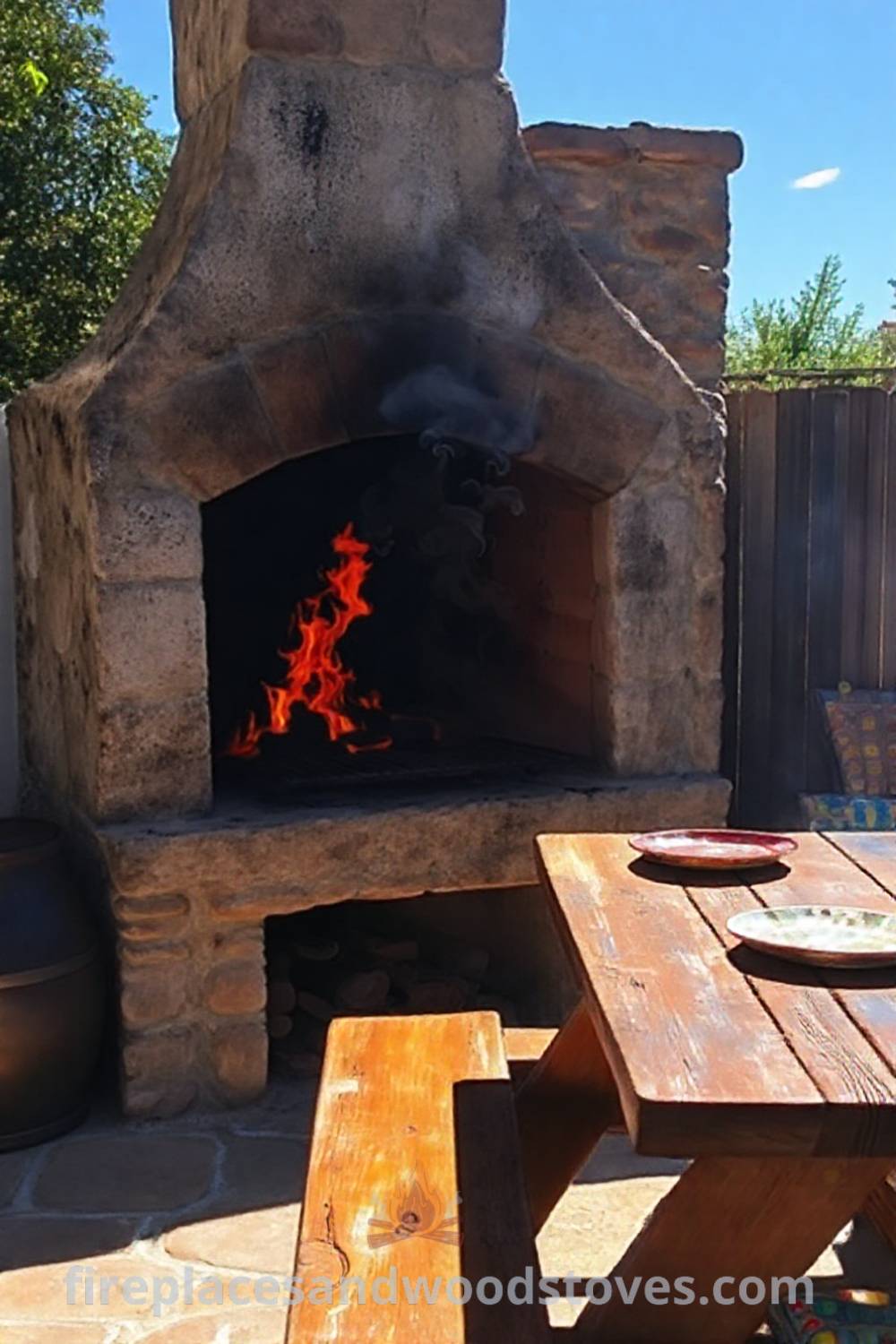 Rustic fireplace BBQ on a sun-drenched patio with a weathered stone structure and vibrant flames, complemented by a wooden picnic table with mismatched plates and colorful cushions, creating a cozy outdoor atmosphere ideal for memorable gatherings. Visit patioandhomefurniture.com for incredible outdoor design inspiration.