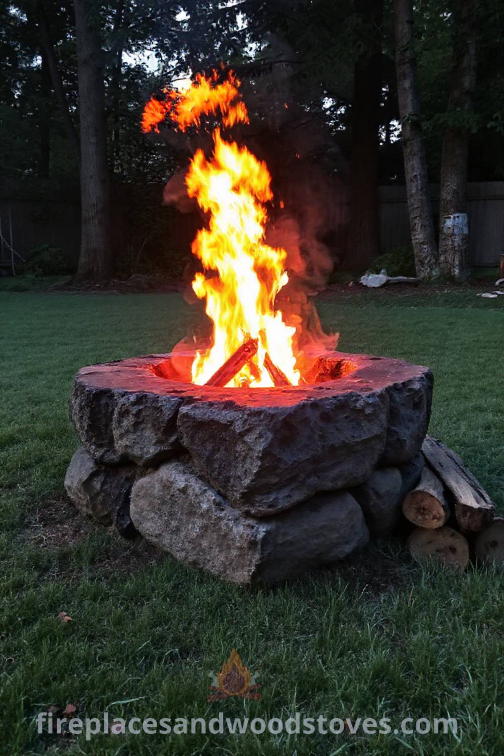 Stone fire pit in a grassy backyard surrounded by trees, with flames crackling and flames casting warm light, creating a cozy atmosphere for gatherings. Visit patioandhomefurniture.com for stylish outdoor design ideas that will wow your guests.