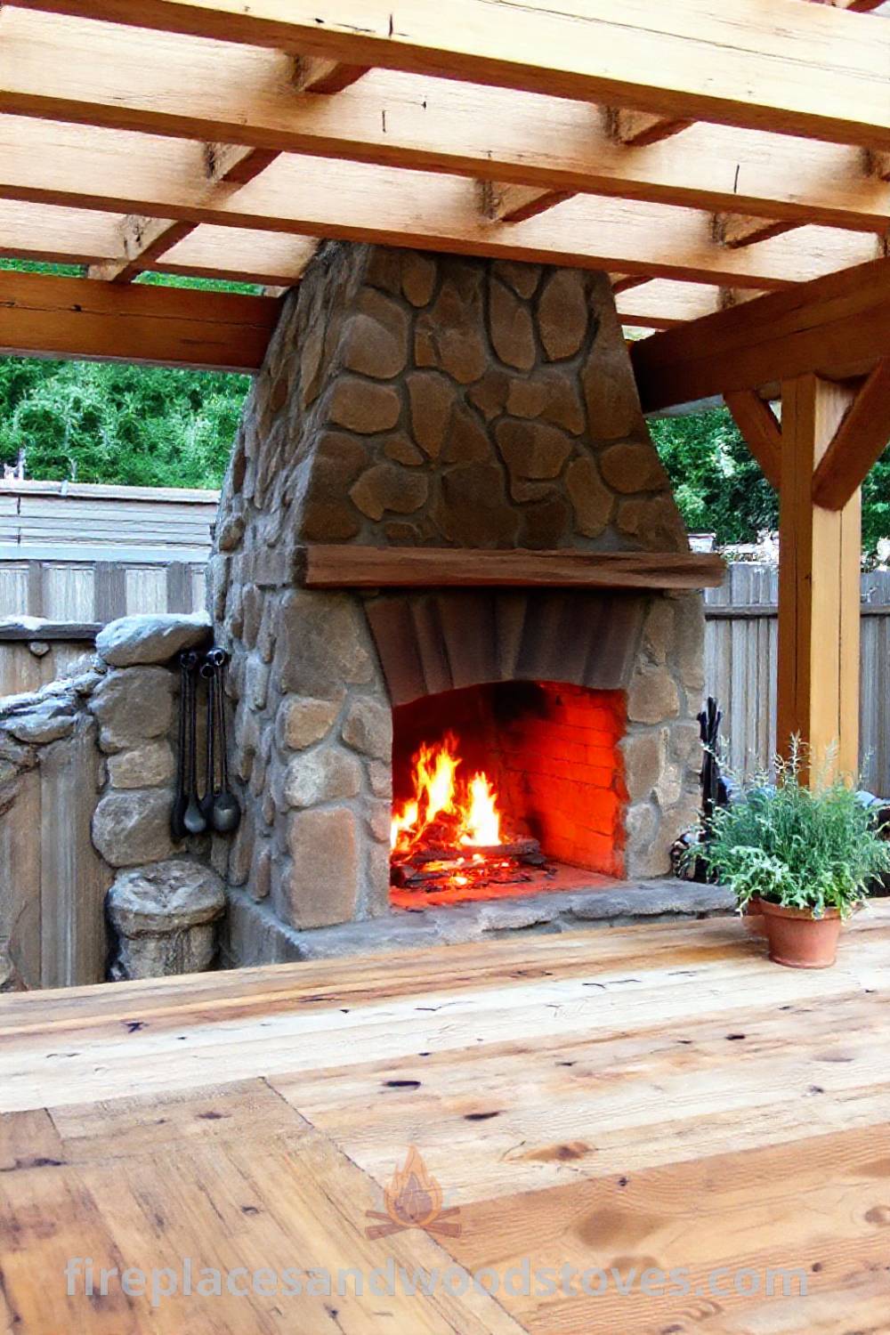 Outdoor kitchen with a rustic wood-burning fireplace under a wooden pergola, complemented by a reclaimed wood counter and hanging utensils, creating a cozy and stylish atmosphere. Visit patioandhomefurniture.com for incredible ideas and inspiration to transform your backyard.