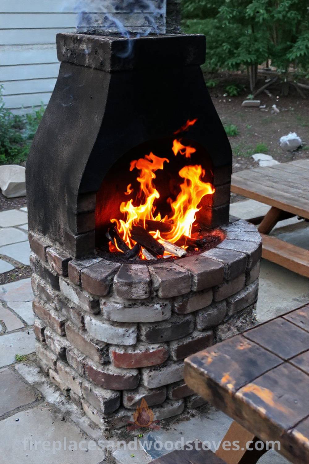 Rustic fireplace BBQ on a stone patio with vibrant flames, a wooden picnic table nearby, and an inviting atmosphere for a cozy evening under the stars. Visit patioandhomefurniture.com for incredible outdoor design ideas that will wow your guests.