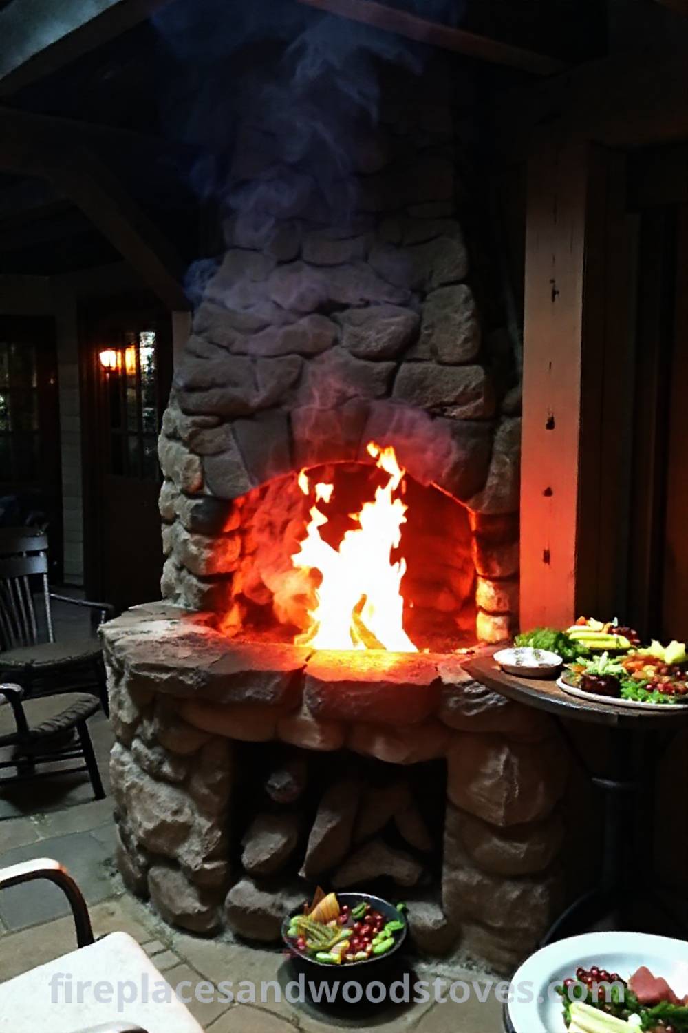 Stone fireplace BBQ in a quaint outdoor patio, featuring flickering flames, rugged surfaces, and a table with fresh vegetables, creating a cozy and inviting atmosphere for gatherings. Visit patioandhomefurniture.com for stylish outdoor design ideas that will wow your guests.