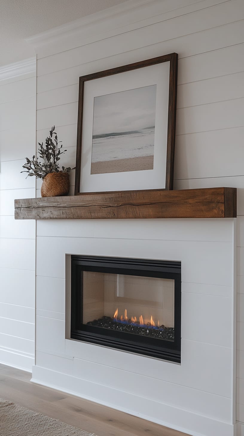 An elegant beach house fireplace with white shiplap walls, a wooden shelf holding framed art, and a cozy ambiance created by a burning fire.