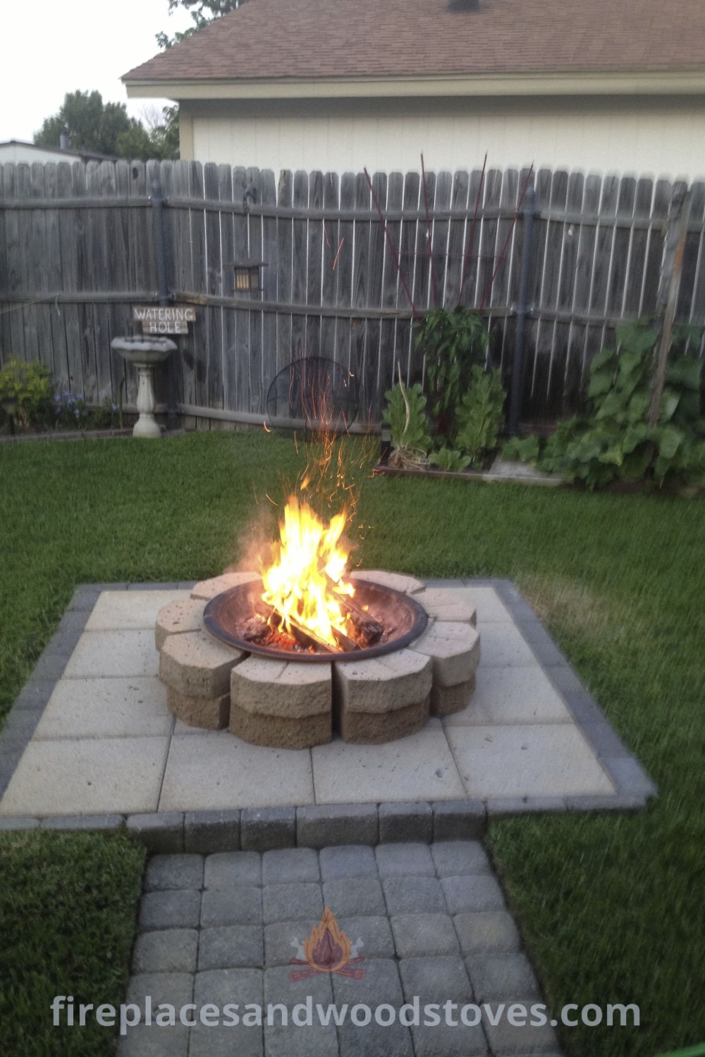 A paver fire pit in a yard, surrounded by grass and a fence, showcasing stylish design ideas for outdoor spaces. Perfect for cozy gatherings and enhancing your patio area. Discover more fire pit inspiration at patioandhomefurniture.com.