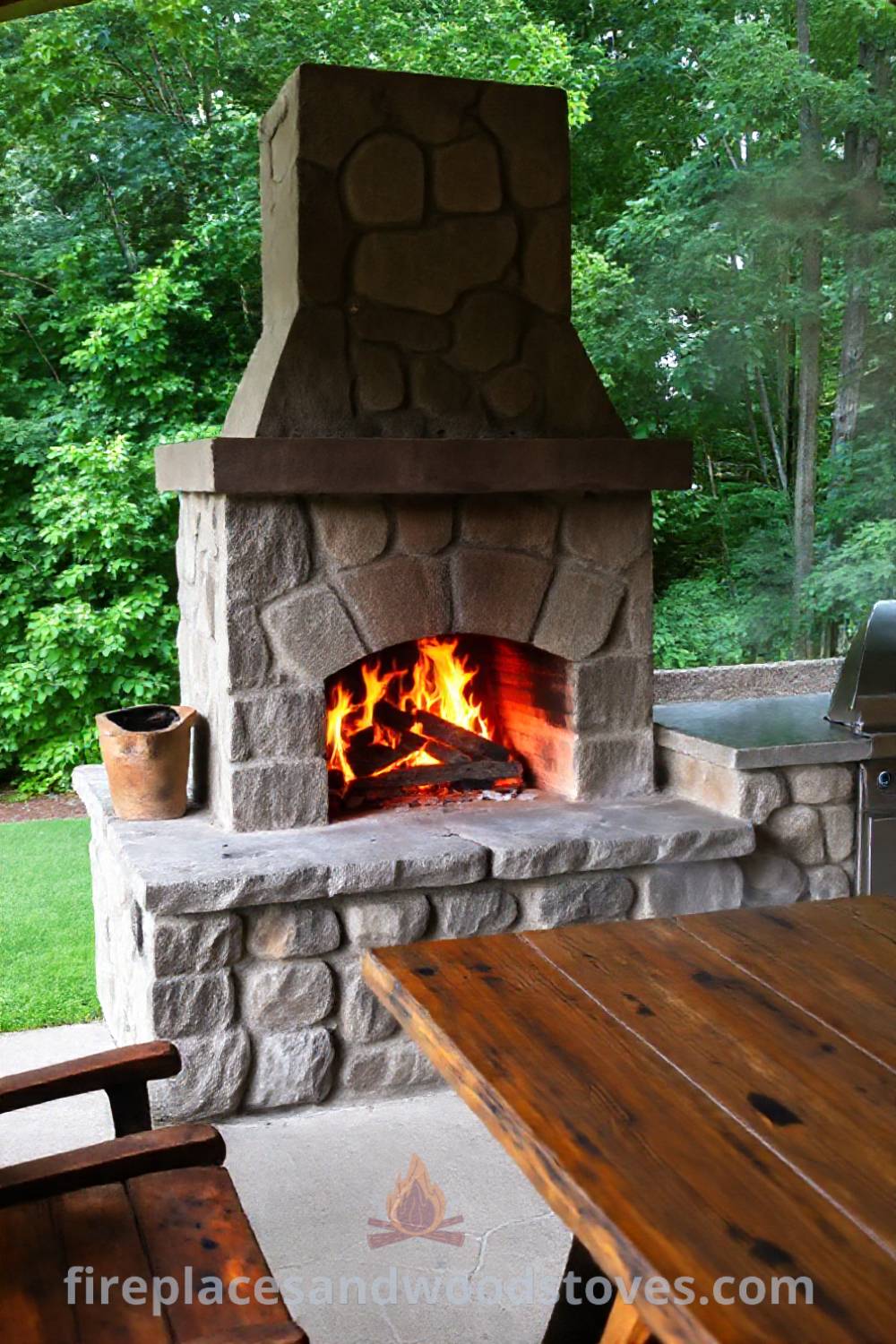 Outdoor kitchen featuring a cozy wood-burning fireplace surrounded by greenery, with a rustic stone hearth and wooden table, creating an inviting space for gatherings and delicious grilled food. Visit patioandhomefurniture.com for stylish outdoor design ideas that will wow your guests.