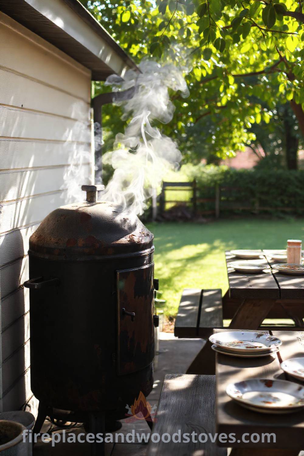 A cozy patio featuring a sturdy barbecue smoker, weathered picnic table with mismatched plates, and dappled sunlight filtering through tree leaves, creating an inviting outdoor space for summer gatherings. Visit patioandhomefurniture.com for stylish outdoor design ideas that will wow your guests.
