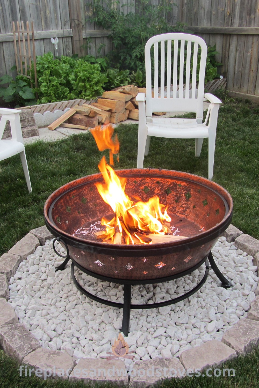 A fire pit in the middle of a yard with white chairs around it and a wooden fence behind it, featuring pebbles for decor. This setup offers portable fire pit ideas that elevate your backyard experience. Discover more stylish outdoor inspiration at patioandhomefurniture.com.