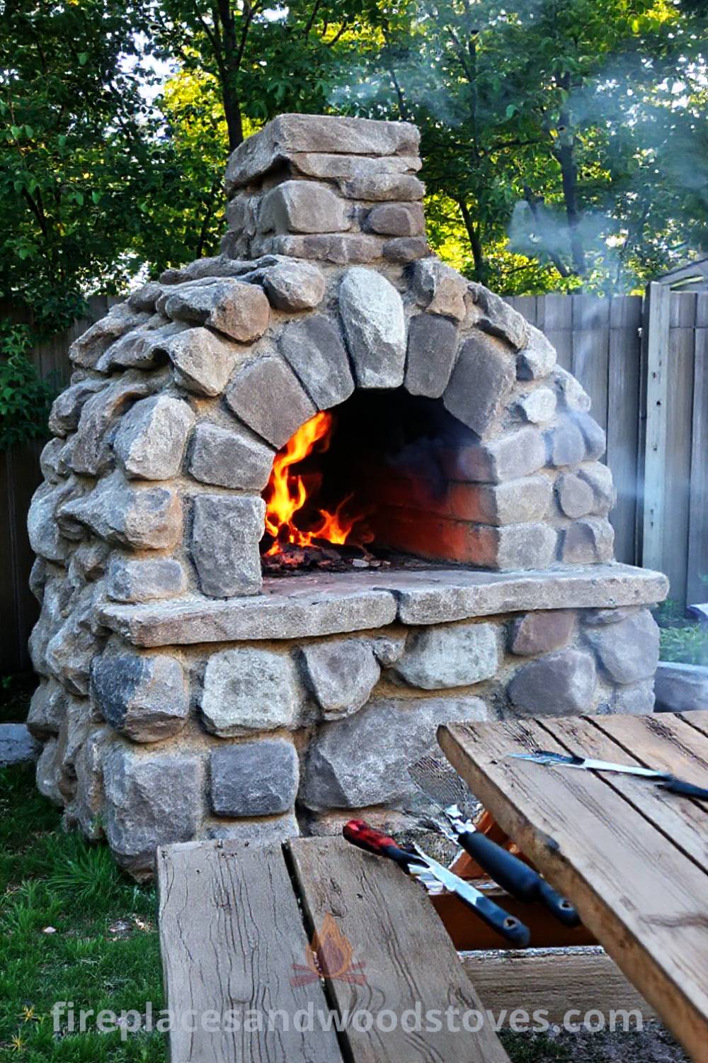 Rugged stone BBQ oven in a sun-dappled backyard with a wooden picnic table set for a casual meal, surrounded by warm firelight and an inviting atmosphere, perfect for summer gatherings. Visit patioandhomefurniture.com for incredible ideas to transform your backyard.