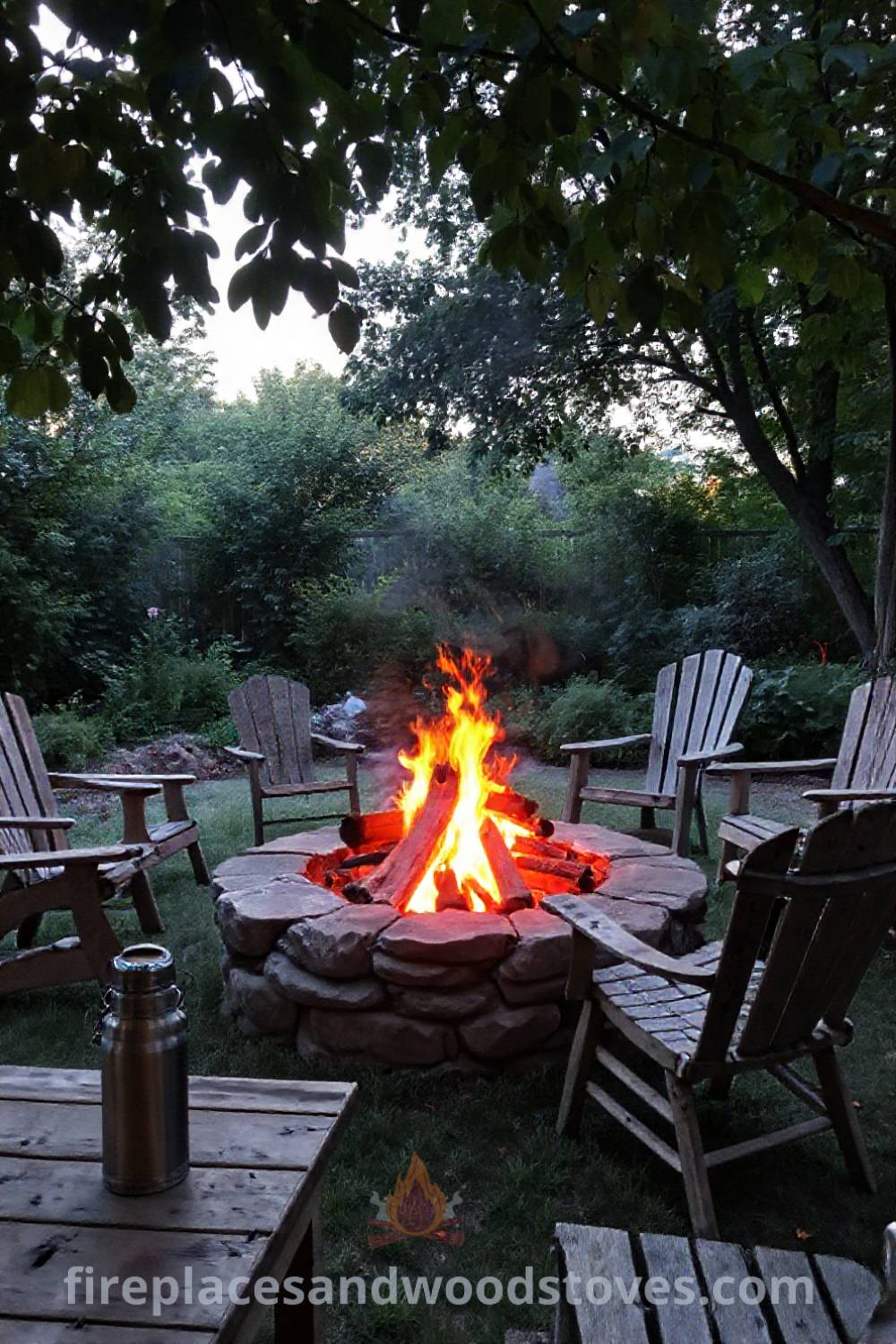 Stone fire pit in a lush garden surrounded by mismatched outdoor chairs, with logs smoldering and a nearby wooden table holding a thermos; a perfect cozy gathering spot for outdoor inspiration. Visit patioandhomefurniture.com for incredible ideas that will wow your guests.