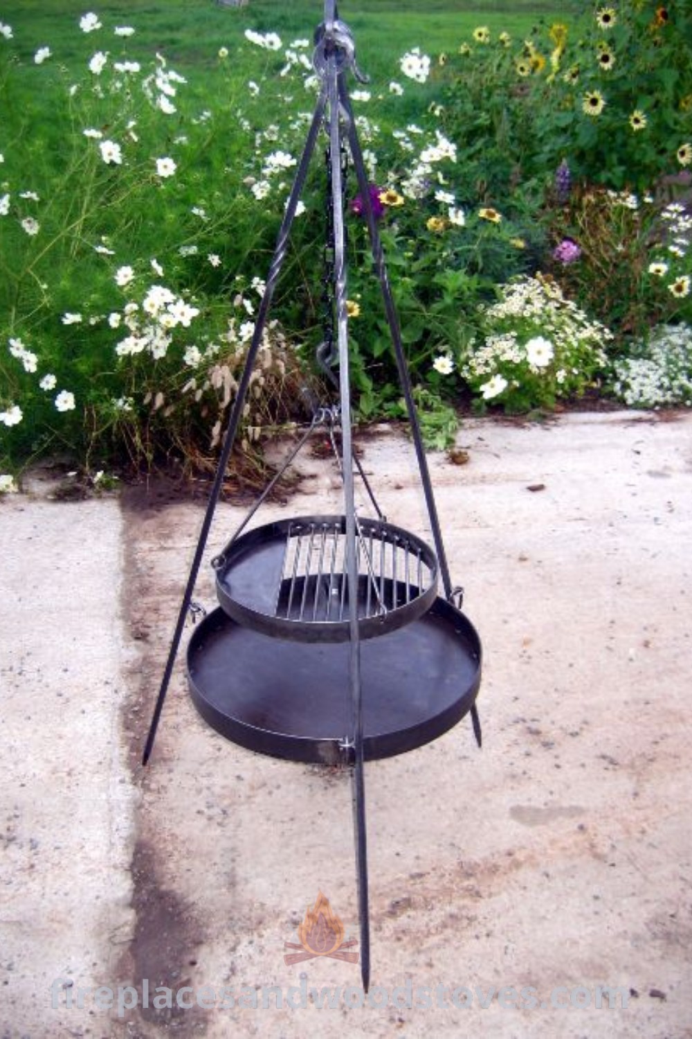 An outdoor fire pit on a cement slab surrounded by flowers and grass, featuring stylish decorative elements and functional outdoor cooking options, perfect for backyard gatherings. Discover more design ideas at patioandhomefurniture.com.