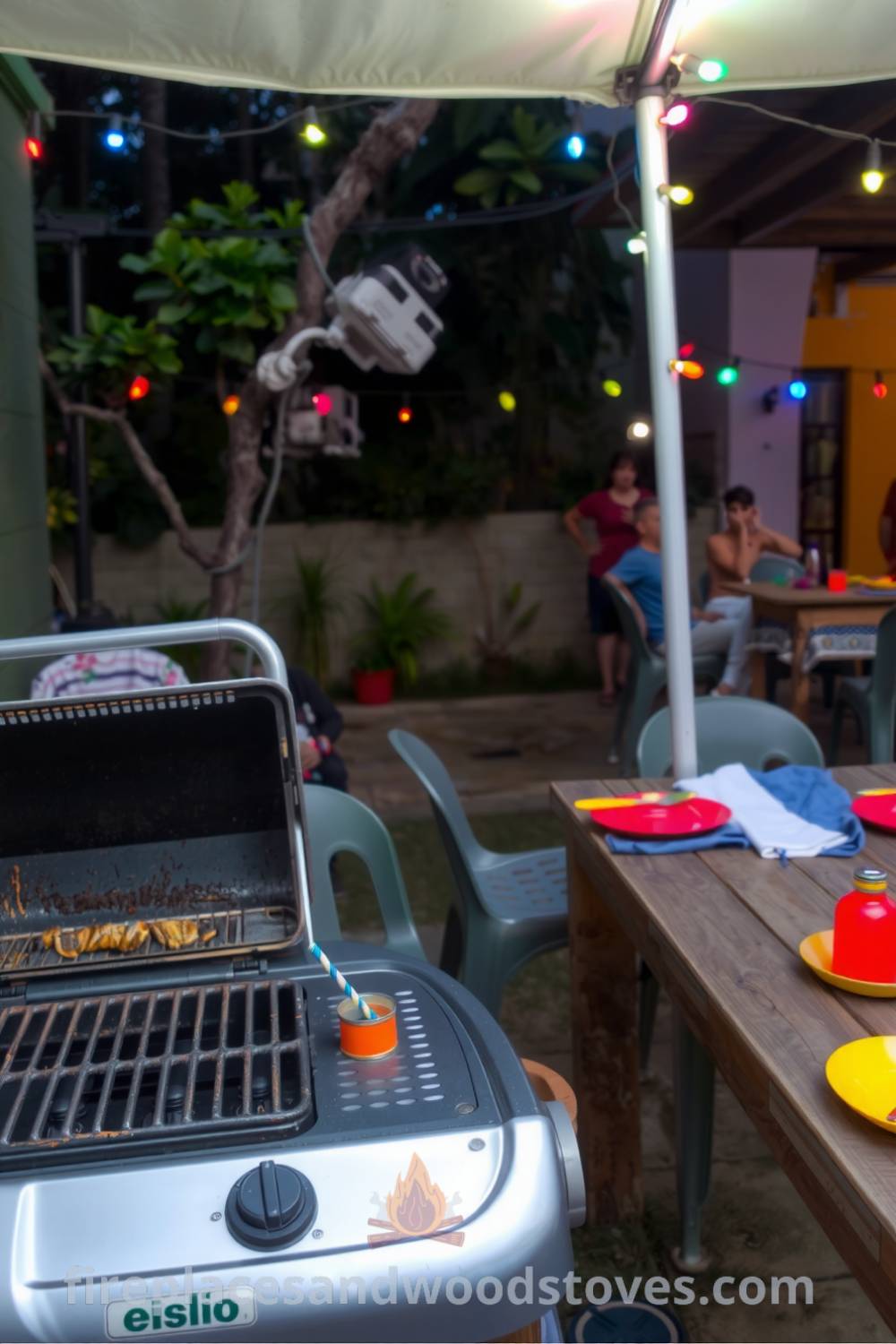 Cozy backyard barbecue setup featuring a small gas grill under a canopy, charred grill grates, a weathered wooden table with colorful plates and utensils, and twinkling string lights, creating an inviting atmosphere for gatherings at fireplacesandwoodstoves.com.