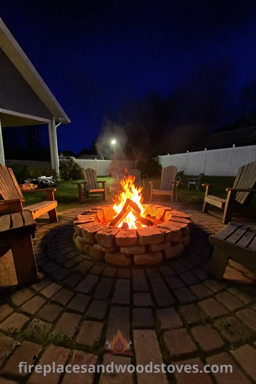 Stone fire pit in a backyard setting surrounded by worn wooden benches and lawn chairs, illuminated by warm flames, creating a cozy and inviting atmosphere. Visit patioandhomefurniture.com for incredible ideas that will wow your guests and transform your backyard.