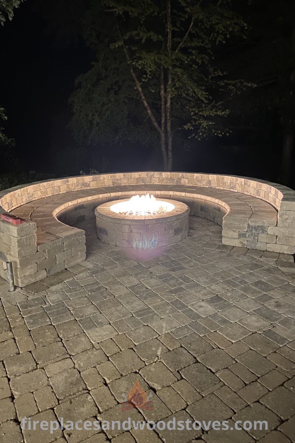 A circular fire pit on a brick patio next to a forest at night, showcasing a round outdoor fire pit area with cozy seating. This design transforms your backyard into a stylish retreat for entertaining guests. Discover more inspiration at patioandhomefurniture.com.