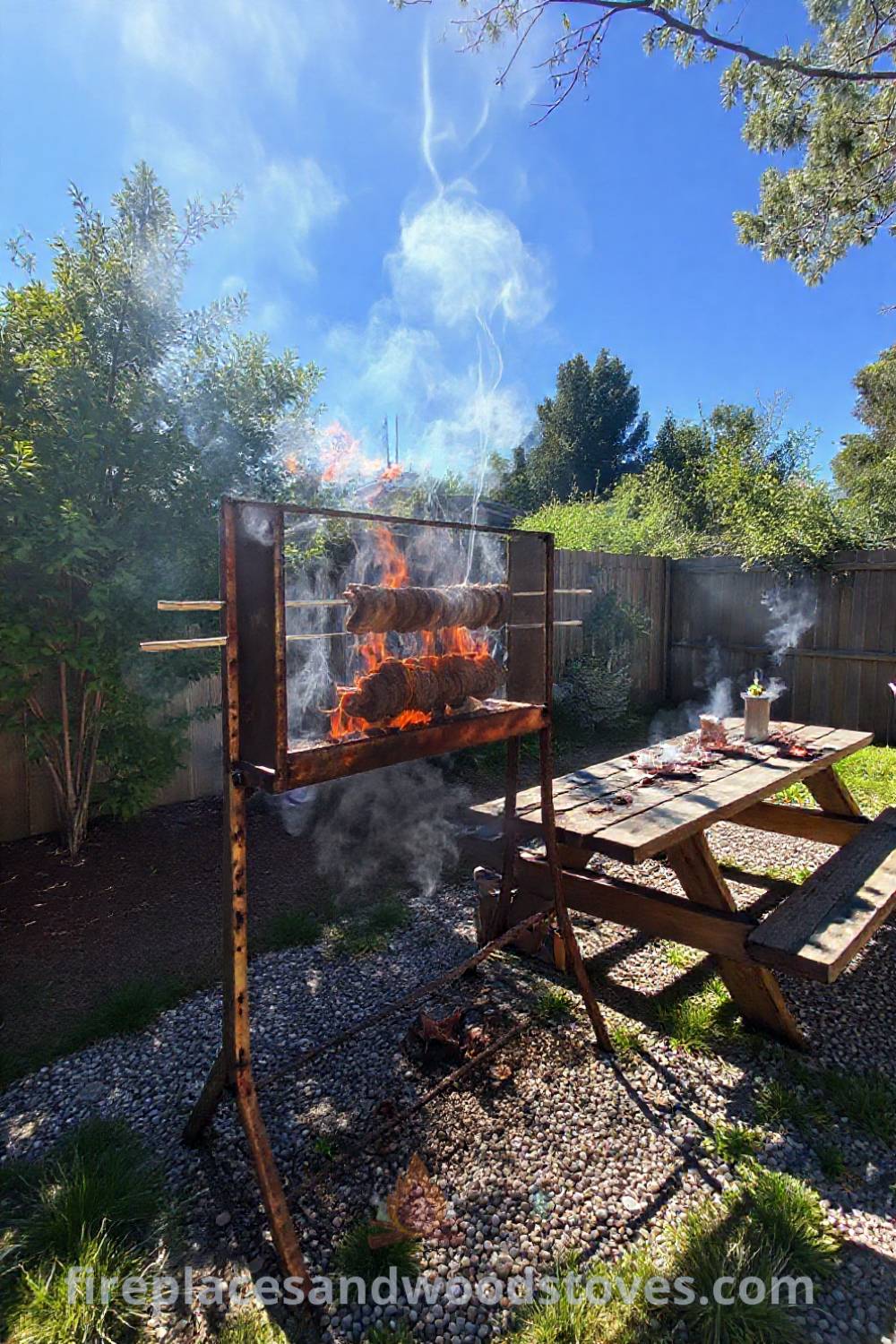 Spit BBQ in a sunlit backyard with a rusted metal frame, flames cooking skewered meat, a wooden picnic table, and patches of grass on gravel, creating a cozy outdoor gathering space. Visit patioandhomefurniture.com for incredible outdoor design inspiration.