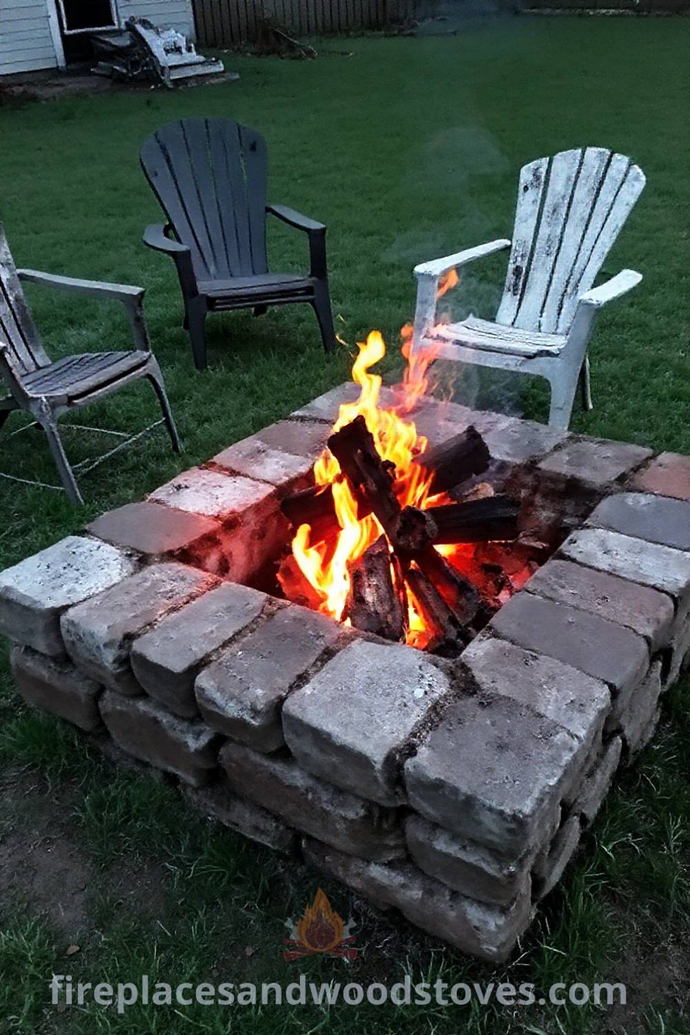 Stone fire pit in a grassy backyard with flickering flames, mismatched chairs, and gathered friends, creating a cozy and inviting atmosphere for memorable gatherings. Visit patioandhomefurniture.com for creative outdoor ideas that will wow your guests.
