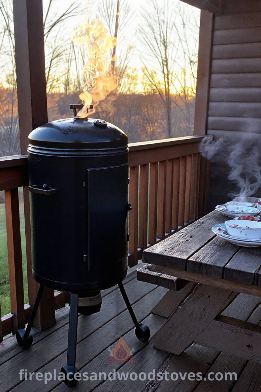 Vertical smoker barbecue on a wooden deck with picnic table and mismatched dishes, surrounded by golden evening light and swirling smoke, creating a cozy atmosphere for outdoor gatherings. Visit patioandhomefurniture.com for stylish outdoor design ideas that will wow your guests.