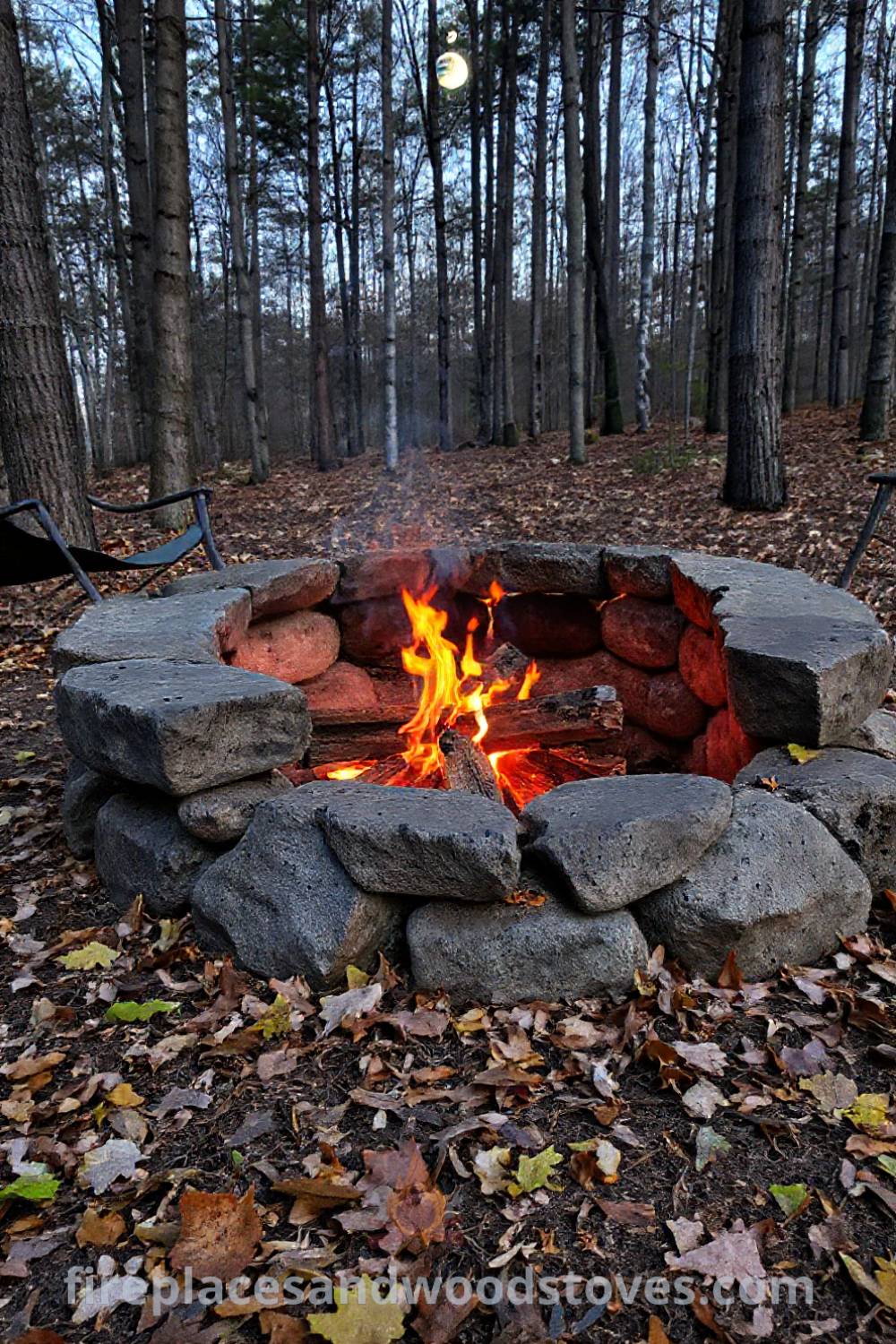 Rustic stone camping fire pit surrounded by trees and fallen leaves, with flickering flames and camp chairs creating a cozy outdoor setting. Visit patioandhomefurniture.com for stylish design ideas that will wow your guests.