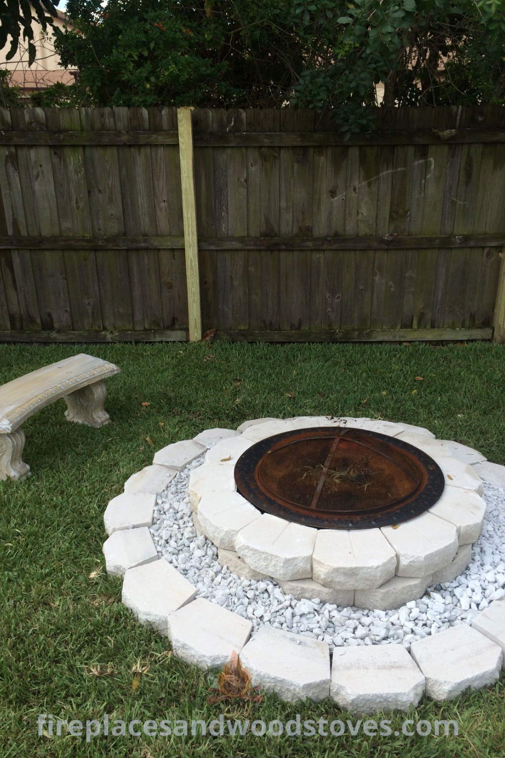 A fire pit in a beautiful yard surrounded by benches and a fence, featuring elegant white rocks and stone designs for a stylish outdoor gathering. Discover more incredible ideas for transforming your backyard at patioandhomefurniture.com.