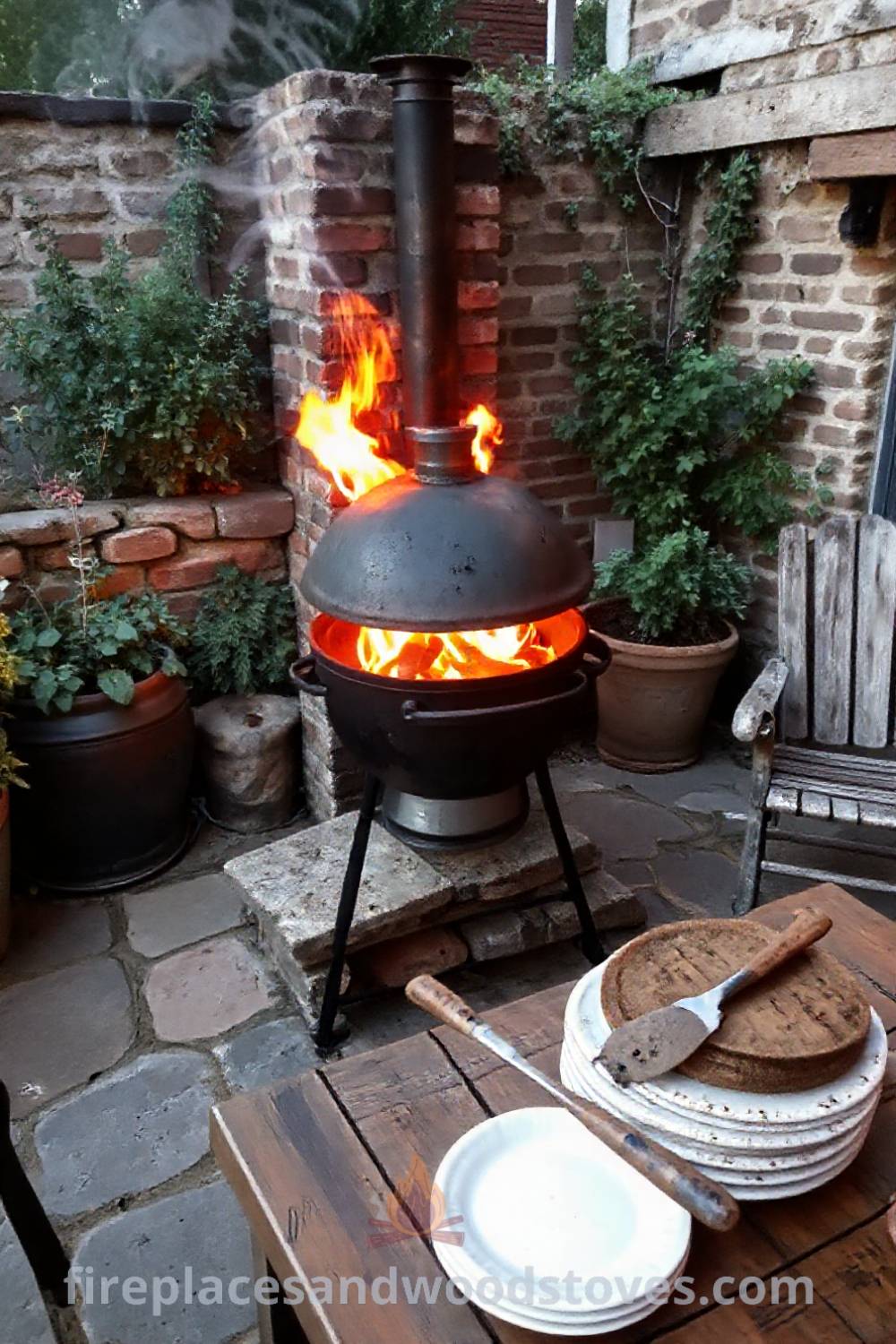 Rustic wood-fired barbecue on a stone patio with gentle smoke, surrounded by plants and weathered outdoor furniture, creating a cozy atmosphere for summer gatherings. Visit patioandhomefurniture.com for stylish outdoor design ideas that will wow your guests.