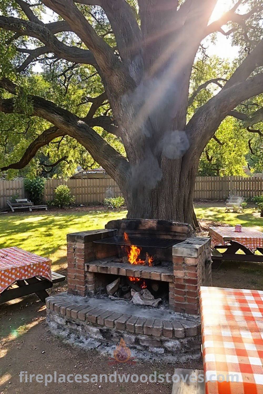 Rustic wood-fired barbecue under an oak tree in a sunlit backyard with weathered brick surround, flames glowing on grates, and picnic tables with checkered cloths, creating a cozy and inviting atmosphere for summer feasts. Visit patioandhomefurniture.com for incredible outdoor design inspiration.