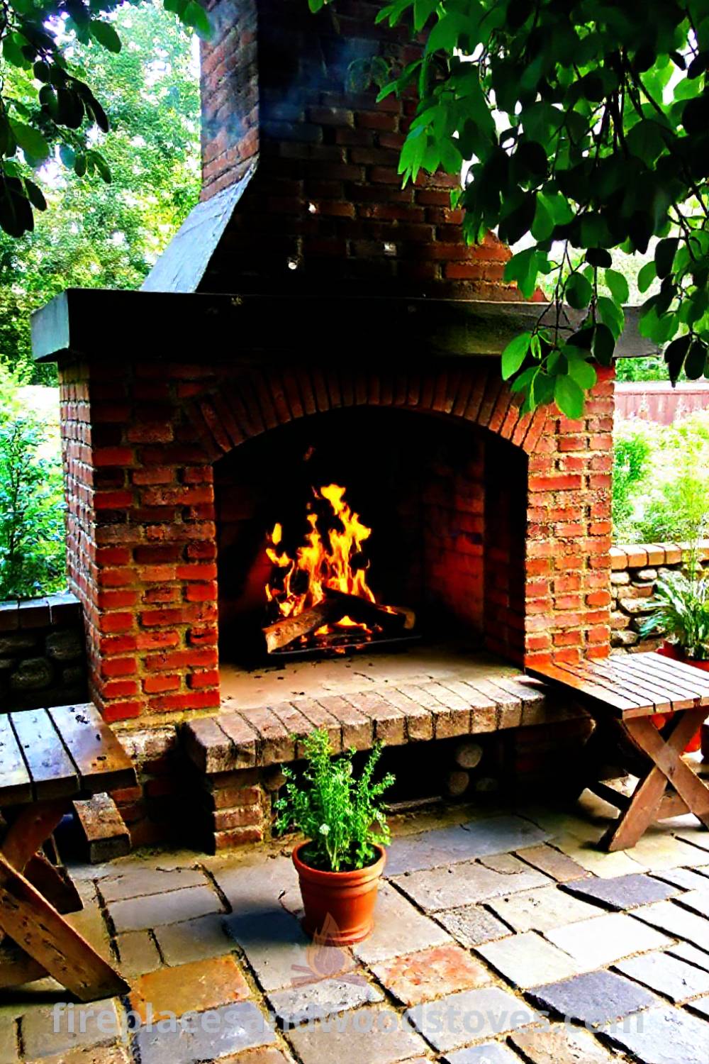 Rustic fireplace BBQ on a stone-paved patio with red-brick surround, flames crackling and wooden tables filled with grilling essentials, and potted herbs enhancing the cozy outdoor atmosphere. Visit patioandhomefurniture.com for stylish designs transforming your backyard into an incredible oasis.