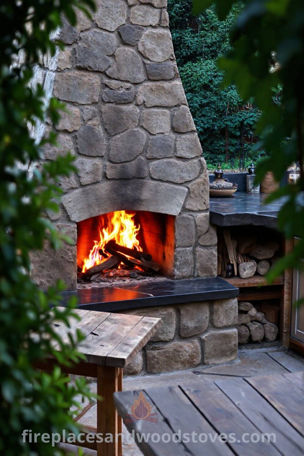 Rustic outdoor kitchen with a wood-burning fireplace made of uneven stone, surrounded by a slate countertop and fresh ingredients, set within lush greenery. Visit patioandhomefurniture.com for incredible ideas that will wow your guests.