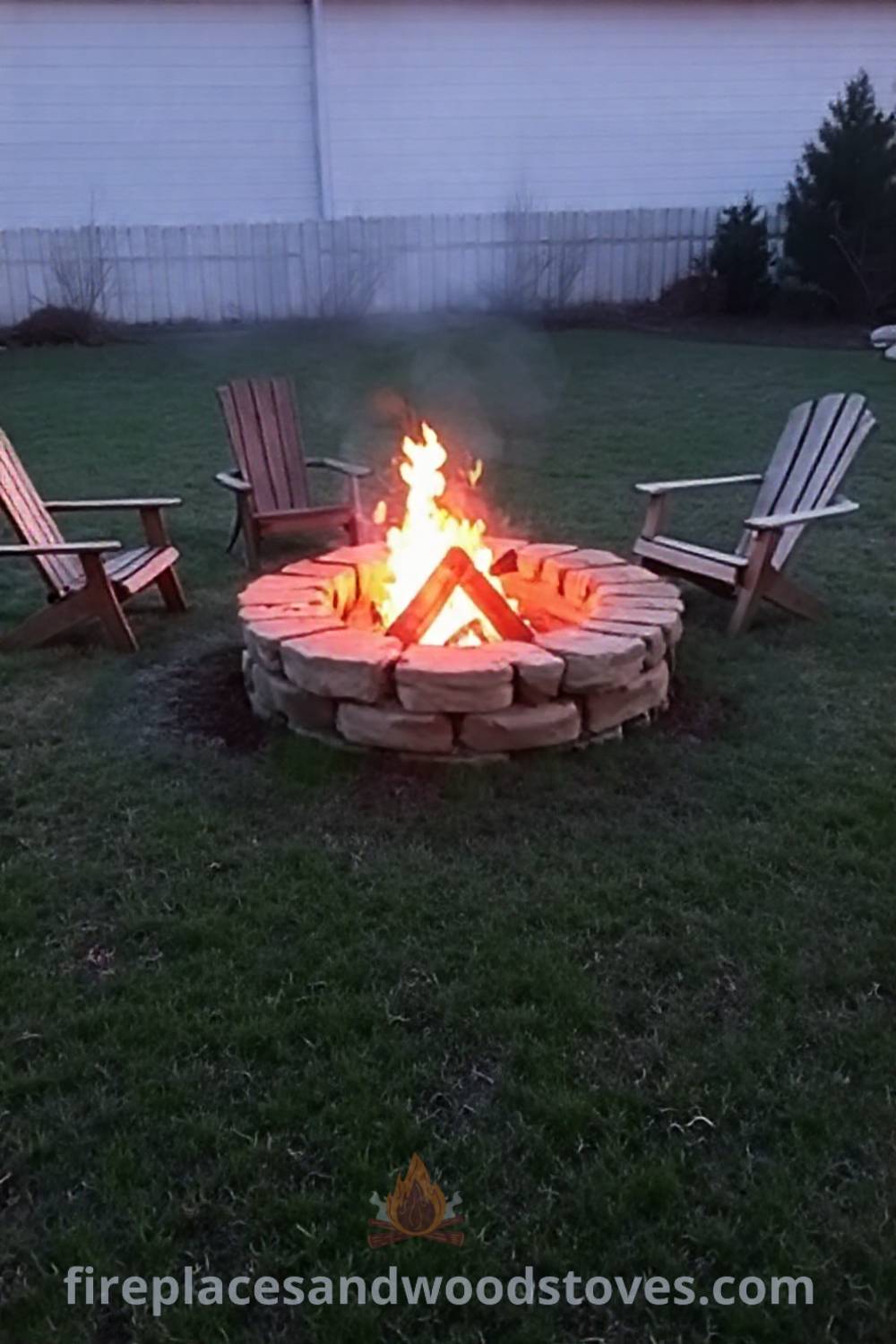 Stone fire pit surrounded by mismatched wooden chairs in a grassy backyard, creating a cozy and inviting atmosphere for gatherings. Visit patioandhomefurniture.com for stylish outdoor design ideas that will wow your guests.