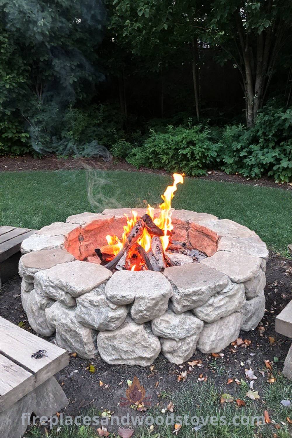 Rustic stone fire pit in a lush backyard, surrounded by wooden seating, with flickering flames creating a warm glow, perfect for cozy gatherings. Visit patioandhomefurniture.com for stylish outdoor design ideas that will wow your guests.