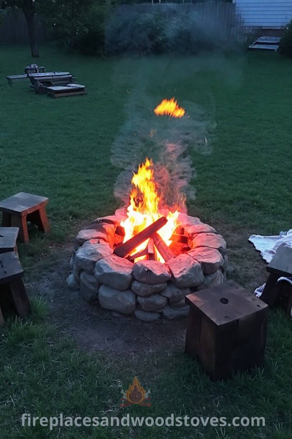 Stone fire pit in a grassy backyard surrounded by wooden stools and blankets, creating a cozy gathering space for warmth and togetherness. Visit patioandhomefurniture.com for inspiration on outdoor designs that will wow your guests.