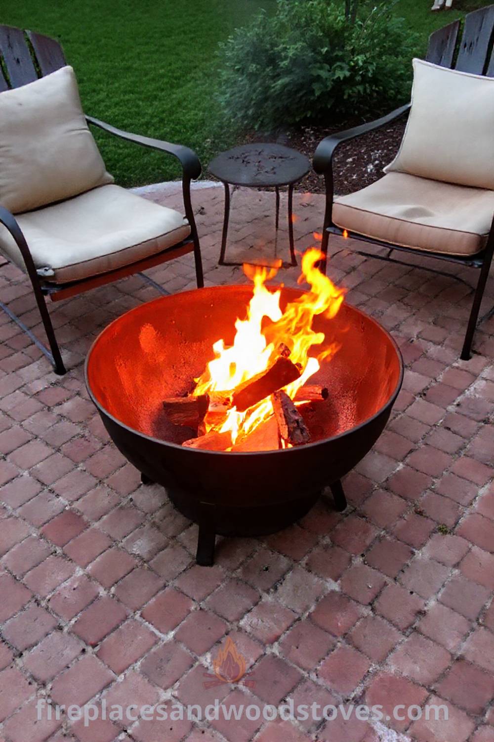 Wood-burning metal fire pit on a brick patio surrounded by mismatched chairs, creating a cozy and inviting outdoor gathering space for friends and family. Get stylish and creative ideas for your backyard at patioandhomefurniture.com.