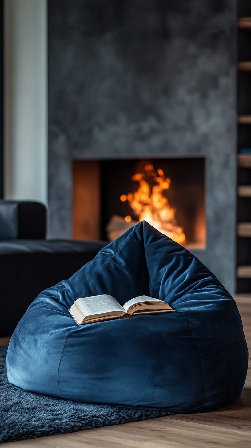 A dark blue velvet bean bag chair positioned in front of a fireplace, with an open book on it, set against a modern minimalist interior with black marble walls.