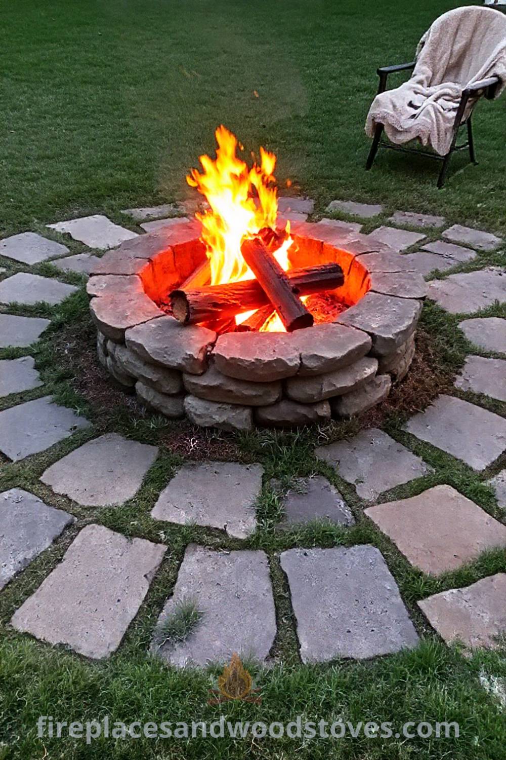 Stone fire pit surrounded by worn paving stones in a grassy backyard, with flickering flames and a cozy blanket on a chair, perfect for inviting warm gatherings. Visit patioandhomefurniture.com for outdoor design inspiration and ideas that will wow your guests.